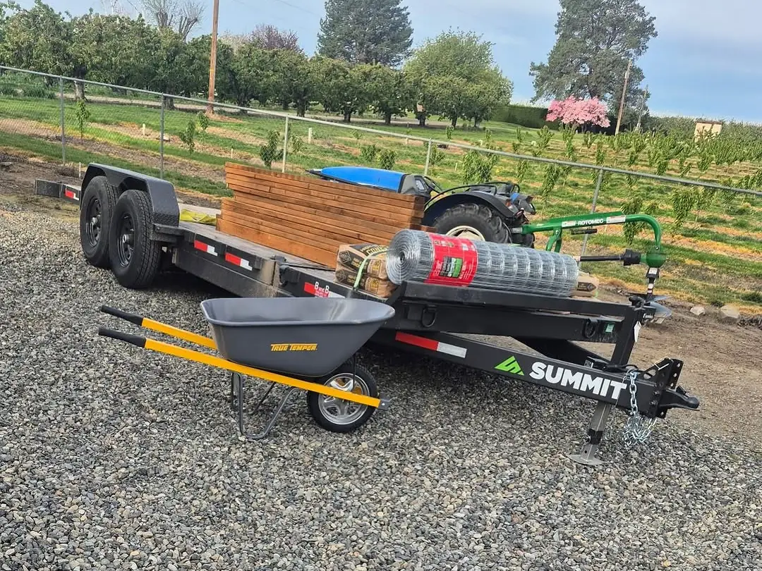 Equipment trailer loaded with lumber, wire and a wheelbarrow beside a compact tractor, general contracting