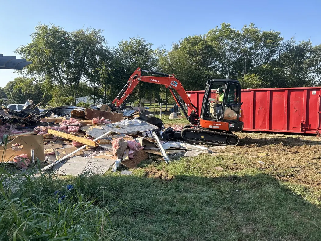 Mini excavator loading wood, metal and insulation debris beside a red roll off dumpster, demolition