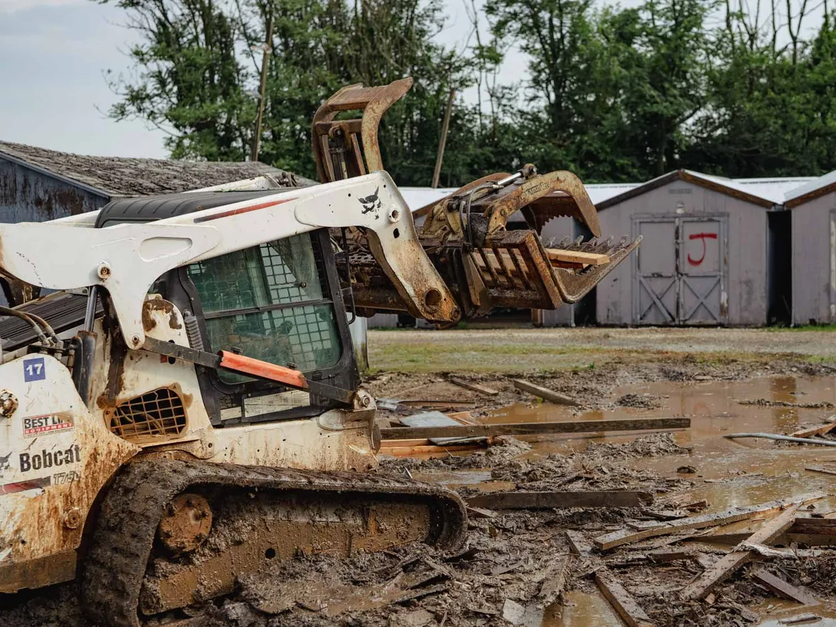 Track loader with a grapple clearing wood debris from a muddy lot beside old sheds, demolition