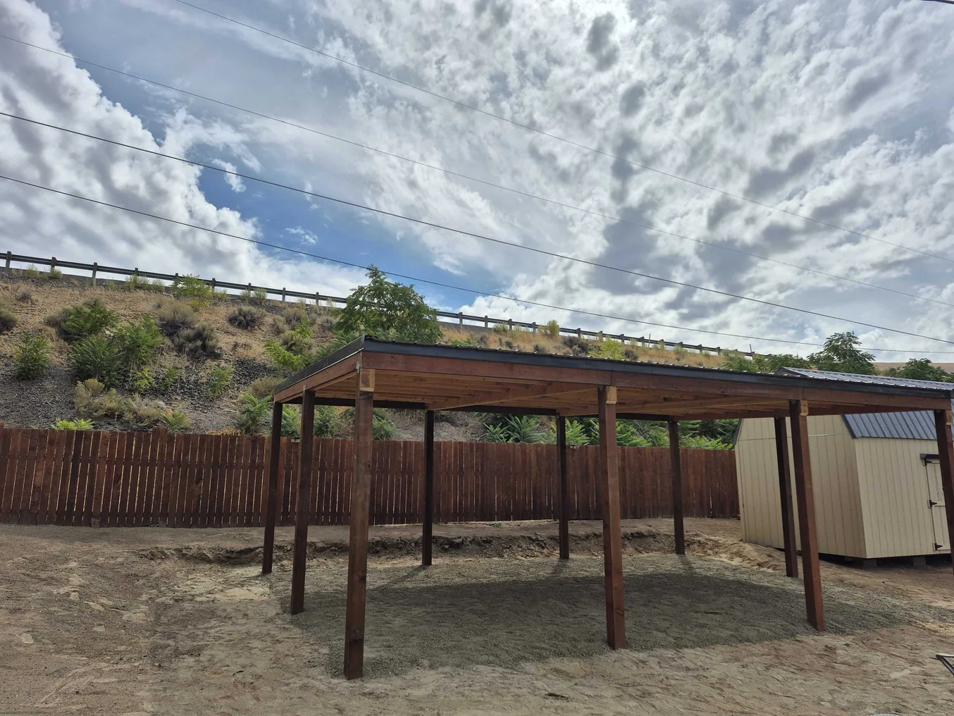 Wood post carport with a metal roof on a gravel pad by a fence, general contracting