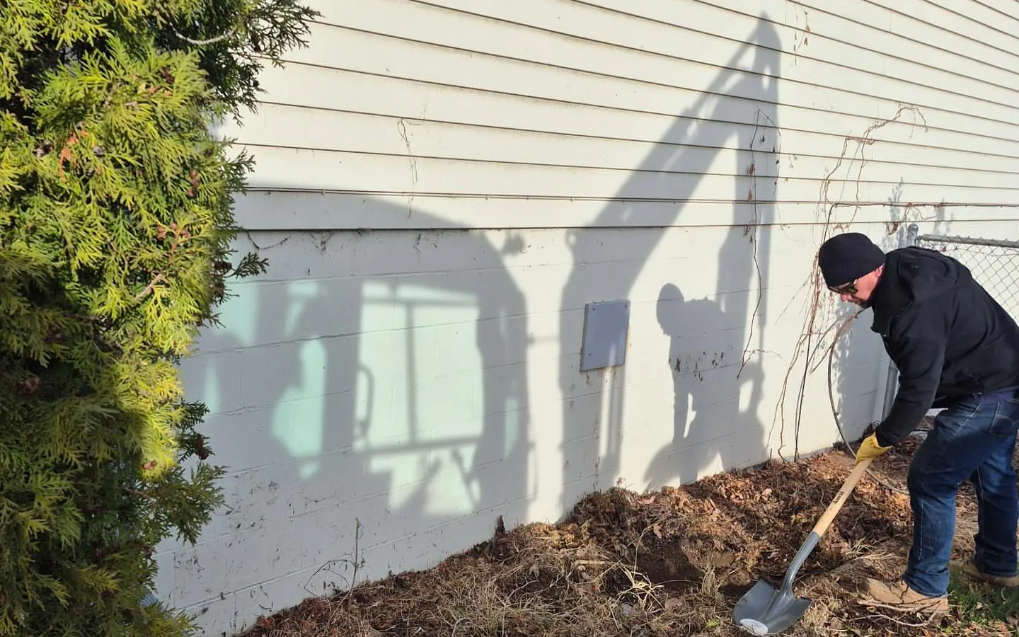 Worker regrading soil along a house foundation with a mini excavator shadow on the wall, grading