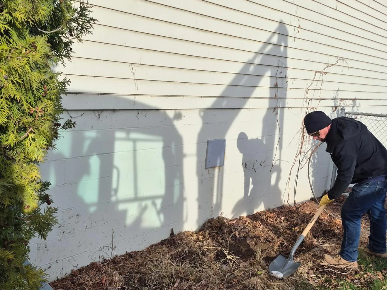 Worker digging along a house wall with the shadow of a mini excavator on the siding, mass excavation