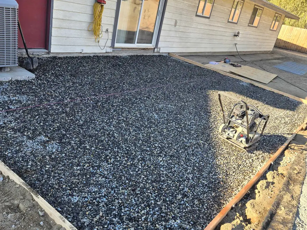 Compacted gravel base inside wood forms with a plate compactor beside a house, site preparation